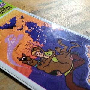 May include: A pack of Scooby-Doo Halloween stickers featuring Scooby and Shaggy in a spooky setting with a haunted house, bats, and a ghost. The stickers are on a purple, orange, and yellow background.