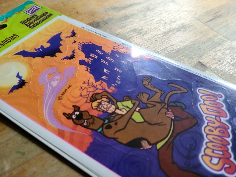 May include: A pack of Scooby-Doo Halloween stickers featuring Scooby and Shaggy in a spooky setting with a haunted house, bats, and a ghost. The stickers are on a purple, orange, and yellow background.
