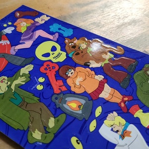 May include: A set of colorful stickers featuring characters from the Scooby-Doo cartoon. The stickers are on a blue background and include a variety of characters, including Scooby-Doo, Shaggy, Daphne, Velma, and Fred. There are also a variety of spooky elements, such as a ghost, a skull, and a key.