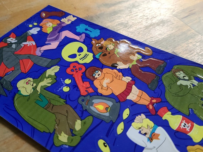 May include: A set of colorful stickers featuring characters from the Scooby-Doo cartoon. The stickers are on a blue background and include a variety of characters, including Scooby-Doo, Shaggy, Daphne, Velma, and Fred. There are also a variety of spooky elements, such as a ghost, a skull, and a key.