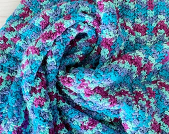 May include: A crocheted blanket in shades of blue, teal, and purple. The blanket is folded and arranged in a spiral shape.