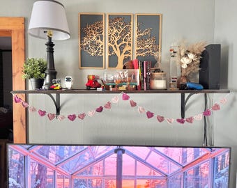 May include: A shelf displays decorative items, including a black lamp, potted plant, books, and a red toy scooter. A crochet heart garland hangs below. Three framed tree art pieces are mounted on the wall above the shelf.