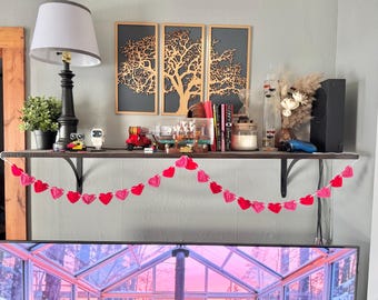 May include: A shelf displays a black lamp, potted plant, decorative items, and books. A red and pink heart garland hangs in front. Three framed tree silhouettes hang on the wall above the shelf.