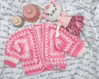 May include: A handmade crochet cardigan featuring a granny square pattern in shades of pink and white, with long sleeves and ribbed cuffs. A plush cat doll wearing a floral dress and a flower crown is positioned next to the cardigan.