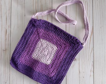 May include: A handmade crochet bag in shades of purple, featuring a square design with a lighter center. The bag has a long, lavender strap for shoulder or crossbody wear. The crochet stitches are visible, adding texture to the bag.