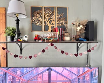 May include: A shelf displays a variety of decorative items, including a black lamp, potted plant, and framed tree art. A crochet heart garland hangs below. Other items include books, candles, and small toys. The color scheme is neutral with pops of red and pink.