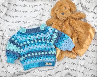 May include: A long-sleeved crochet sweater featuring a multi-colored granny square pattern in shades of navy, royal blue, and light blue, with a small rectangular label at the hem, paired with a plush tan stuffed animal with a satin-like fabric edge.