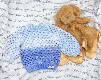 May include: A long-sleeved crochet sweater featuring an ombre color gradient that transitions from light blue at the neckline to a deep royal blue at the hem. The sweater has a textured, open-stitch pattern and a small rectangular label at the bottom.