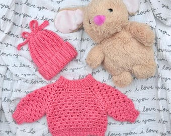 May include: A bright pink crocheted long-sleeve sweater with a ribbed neckline and cuffs, a matching pink crocheted beanie with a bow detail, and a tan plush rabbit toy with a pink nose, all arranged on a white fabric with black text.