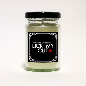 May include: A glass jar candle with a black lid. The candle has a black label with white text that reads "WHEN THE CANDLE IS LIT... LICK MY CLIT" with a red lipstick kiss graphic. The candle is filled with a light beige wax.