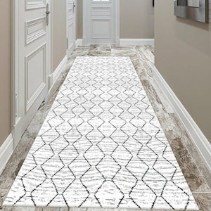Puede incluir: Una alfombra larga y rectangular blanca con un patrón geométrico negro se encuentra sobre un suelo de mármol en un pasillo. El pasillo presenta puertas blancas, molduras y paredes de tonos neutros, creando un espacio luminoso y aireado. El diseño de la alfombra añade un toque moderno.