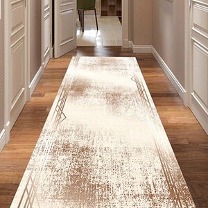 May include: A rectangular hallway rug with a distressed, textured design. The rug features a cream-colored center with brown accents and geometric border details. The rug is placed on a wooden floor.