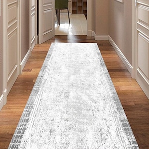 Puede incluir: Una alfombra rectangular de aspecto envejecido en un pasillo. La alfombra presenta una combinación de colores gris claro y blanco con un borde estampado. El pasillo tiene pisos de madera y puertas blancas.