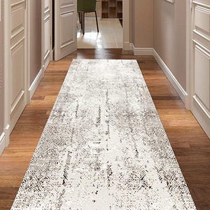 Puede incluir: Una alfombra larga y rectangular con un patrón desgastado en tonos blancos y grises. La alfombra está colocada sobre un suelo de madera en un pasillo, con puertas y paredes blancas a ambos lados. El pasillo conduce a una habitación con una silla verde.