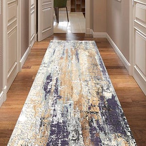 May include: A rectangular rug with an abstract design in beige, grey, and purple tones. The rug is laid in a hallway with wooden flooring and white doors. The rug's dimensions are not visible, but it is likely over 100 cm in length.
