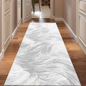 Può includere: Un lungo tappeto rettangolare bianco con un motivo fluente e piumato. Il tappeto è posto su un pavimento in legno in un corridoio, con porte e pareti bianche su entrambi i lati. Il corridoio conduce a una stanza con una sedia verde.