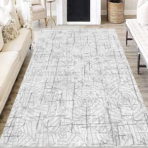 Puede incluir: Una alfombra rectangular con un patrón geométrico en tonos blancos y grises. La alfombra está colocada en un salón, con una chimenea, un sofá y una silla en el fondo. El diseño presenta líneas que se cruzan.