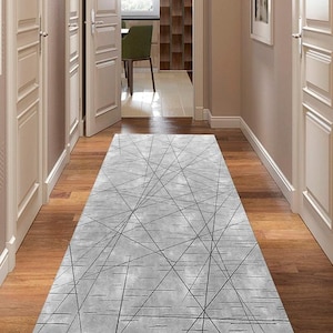 Puede incluir: Una alfombra rectangular gris con un diseño geométrico de líneas negras se encuentra sobre un suelo de madera en un pasillo. El patrón de la alfombra consiste en líneas que se cruzan, creando una estética moderna. El pasillo tiene puertas y paredes blancas.
