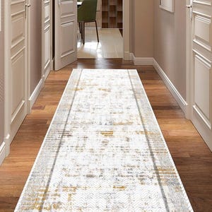 Puede incluir: Una alfombra rectangular para pasillo con un patrón desgastado en tonos blancos, dorados y grises. La alfombra está colocada sobre un suelo de madera, y el pasillo tiene puertas y paredes blancas.