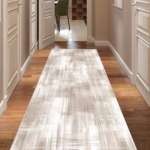 Puede incluir: Una alfombra rectangular de color claro con un patrón geométrico se encuentra sobre un suelo de madera en un pasillo. El diseño de la alfombra presenta una mezcla de líneas grises claras y blancas, creando una apariencia texturizada. El pasillo tiene puertas y paredes blancas.