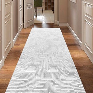 Könnte beinhalten: Ein rechteckiger weißer Teppich mit einem geometrischen Muster aus geraden Linien, der auf einem Holzboden in einem Flur liegt. Das Design des Teppichs erzeugt ein strukturiertes Aussehen. Der Flur hat weiße Türen und Wände.