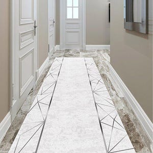 Puede incluir: Una alfombra de pasillo blanca con un diseño geométrico. La alfombra es principalmente blanca con una apariencia texturizada y presenta un borde de líneas geométricas plateadas. El pasillo tiene puertas blancas y paredes de color claro.