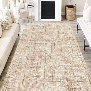 Puede incluir: Alfombra rectangular beige con un patrón geométrico de líneas entrecruzadas. La alfombra está colocada en un salón, con una chimenea blanca, un sofá y un sillón en el fondo. El diseño de la alfombra añade textura y calidez al espacio.