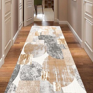 Puede incluir: Una alfombra rectangular para pasillo con un patrón geométrico en tonos beige, gris y marrón. La alfombra presenta grandes formas circulares y rectangulares superpuestas. La alfombra está colocada sobre un suelo de madera, y el pasillo está enmarcado por puertas y paredes blancas.