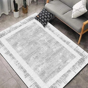 Puede incluir: Una alfombra rectangular con un diseño desgastado y texturizado en tonos grises y blancos. La alfombra tiene un borde con un patrón geométrico. La alfombra está colocada sobre un suelo gris, con un sofá gris y cojines decorativos en el fondo.