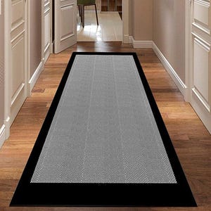 Może przedstawiać: Prostokątny szaro-czarny dywan z wzorem w jodełkę. Dywan ma czarną obwódkę i jest umieszczony na drewnianej podłodze w korytarzu. Korytarz ma białe drzwi i ściany.