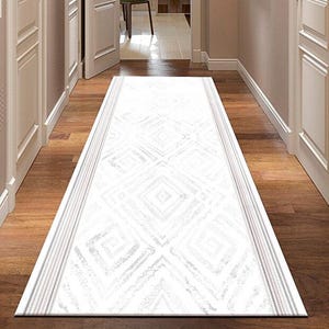 Puede incluir: Una alfombra de pasillo blanca con un diseño geométrico. La alfombra es rectangular y tiene un borde con rayas grises. El suelo es de madera y las paredes están pintadas en un color neutro.