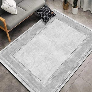 Puede incluir: Una alfombra rectangular con un centro gris claro y un borde gris más oscuro. La alfombra tiene un aspecto texturizado y está colocada sobre un suelo embaldosado. Un sofá gris y cojines decorativos son visibles en el fondo.