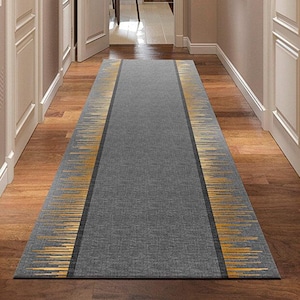 Puede incluir: Una alfombra gris para pasillo con borde dorado y negro. La alfombra es rectangular y está colocada en un pasillo con suelos de madera y puertas blancas. El borde dorado tiene un diseño a rayas.