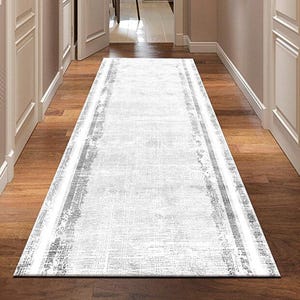 Puede incluir: Una alfombra rectangular blanca para pasillo con un diseño texturizado desgastado y borde gris. La alfombra está colocada sobre un suelo de madera, que conduce a una puerta abierta. El pasillo está flanqueado por puertas blancas y paredes beige.