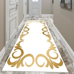 Puede incluir: Una alfombra blanca para pasillo con un diseño dorado ornamentado. La alfombra está centrada en un pasillo con puertas y molduras blancas y una pared gris claro. El suelo es un azulejo tipo mármol.