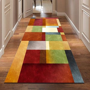 Puede incluir: Una alfombra rectangular con un patrón geométrico en un pasillo. La alfombra presenta un diseño de patchwork con bloques de color rojo, naranja, amarillo, verde y gris. El pasillo tiene pisos de madera y puertas blancas.