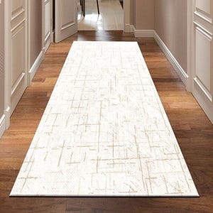 Puede incluir: Una alfombra de pasillo rectangular con fondo crema y un sutil patrón texturizado en beige claro. La alfombra está sobre un suelo de madera oscura, conduciendo a una puerta abierta. Medidas en centímetros.