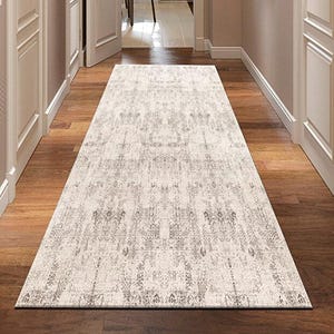 Puede incluir: Una alfombra rectangular de color blanquecino con un diseño desgastado y estampado se encuentra sobre un suelo de madera en un pasillo. El patrón de la alfombra presenta tonos grises claros y oscuros, creando una apariencia texturizada. El pasillo tiene puertas blancas y paredes claras.