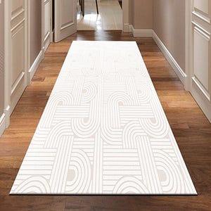 Puede incluir: Una alfombra de pasillo rectangular con un patrón geométrico en blanco y beige. La alfombra está sobre un suelo de madera. El pasillo tiene puertas blancas y paredes beige.