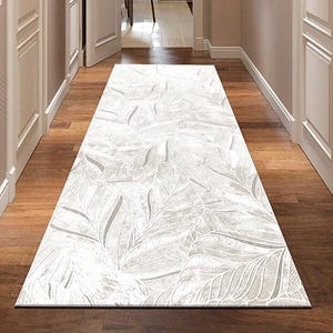 Può includere: Un tappeto rettangolare bianco sporco con un motivo a foglie ripetuto in tonalità di grigio e bianco. Il tappeto è posto in un corridoio con pavimento in legno e porte bianche. Il corridoio conduce a una stanza con una sedia verde.