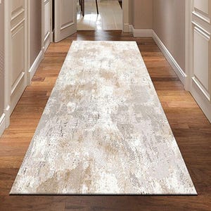 Puede incluir: Una alfombra rectangular con un patrón desgastado en tonos beige, gris y blanco. La alfombra está colocada sobre un suelo de madera en un pasillo, con puertas y paredes blancas.