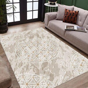 Puede incluir: Alfombra beige con un patrón geométrico blanco y dorado. La alfombra presenta un diseño circular grande con un patrón de flor de la vida, y está colocada sobre un suelo de madera. La alfombra es rectangular y tiene un aspecto envejecido.