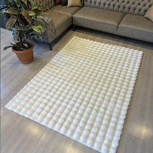 May include: A rectangular, off-white rug with a textured, tufted pattern. The rug is placed on a light-colored wooden floor, with a sectional sofa and potted plant in the background. The rug's dimensions are approximately 60 inches by 80 inches.