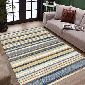 Puede incluir: Una alfombra rectangular con un patrón de rayas en tonos de azul, amarillo, naranja y crema. La alfombra presenta un diseño de borde geométrico en un extremo. La alfombra está colocada sobre un suelo de madera en un salón.