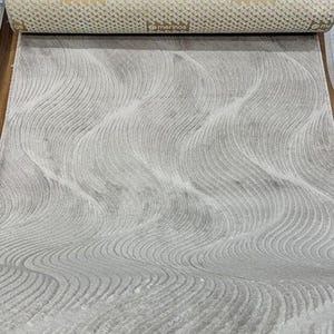 Könnte beinhalten: Eine aufgerollte Teppichrolle mit hellbeigem Rücken und einer grauen, wellenförmigen Oberfläche. Auf der Rückseite ist das Wort "merinos" an mehreren Stellen aufgedruckt. Der Teppich ist ausgerollt und zeigt die graue Musterfläche.