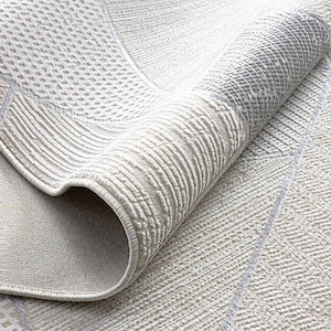 May include: Close-up of a rolled-up area rug with a geometric pattern. The rug features a mix of textures and shades of white, cream, and gray. The design includes lines, squares, and diamond shapes. The rug's edge is visible, showing its thickness and construction.