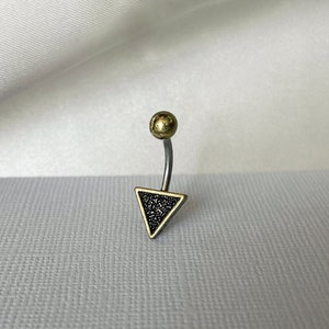 Könnte beinhalten: Ein Bauchnabelpiercing mit einer goldfarbenen Kugel und einem dreieckigen Anhänger. Das Dreieck ist schwarz mit einem strukturierten Muster und mit goldfarbenem Metall umrandet. Der gebogene Steg ist silberfarben.