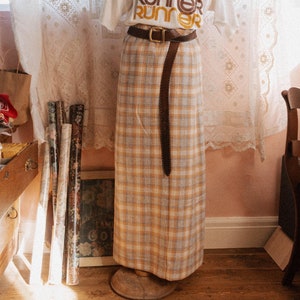 Può includere: Una maglietta bianca con il testo "The Runner" ripetuto quattro volte in un design impilato. Il testo è in nero, marrone e arancione. La maglietta è indossata con una cintura in pelle marrone e una gonna a quadri gialli e blu.