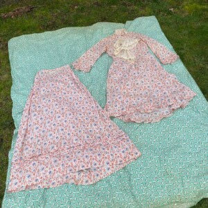 May include: A white dress with a floral print and a lace collar. The dress has long sleeves and a ruffled hem. The dress is laid out on a green and white patterned blanket.