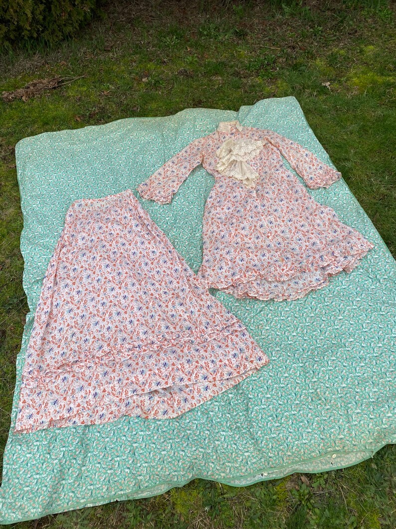 May include: A white dress with a floral print and a lace collar. The dress has long sleeves and a ruffled hem. The dress is laid out on a green and white patterned blanket.